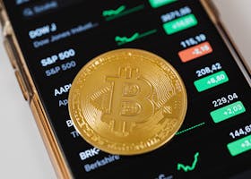A close-up of a Bitcoin coin placed on a mobile device displaying stock market trading data.