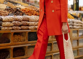 A fashionable woman in a red blazer shops for nuts in a market carrying a mesh bag.